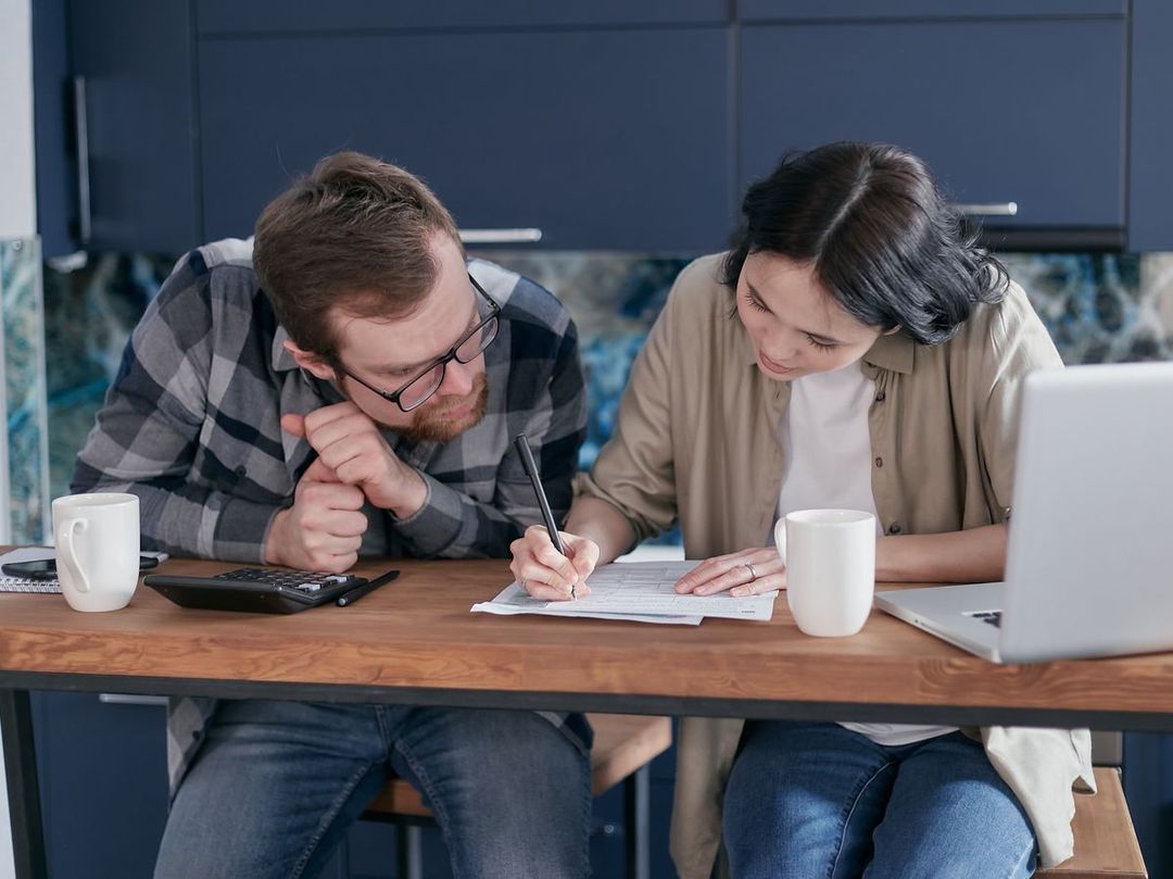 A couple at their kitchen table with a laptop, reviewing their statements together and writing notes